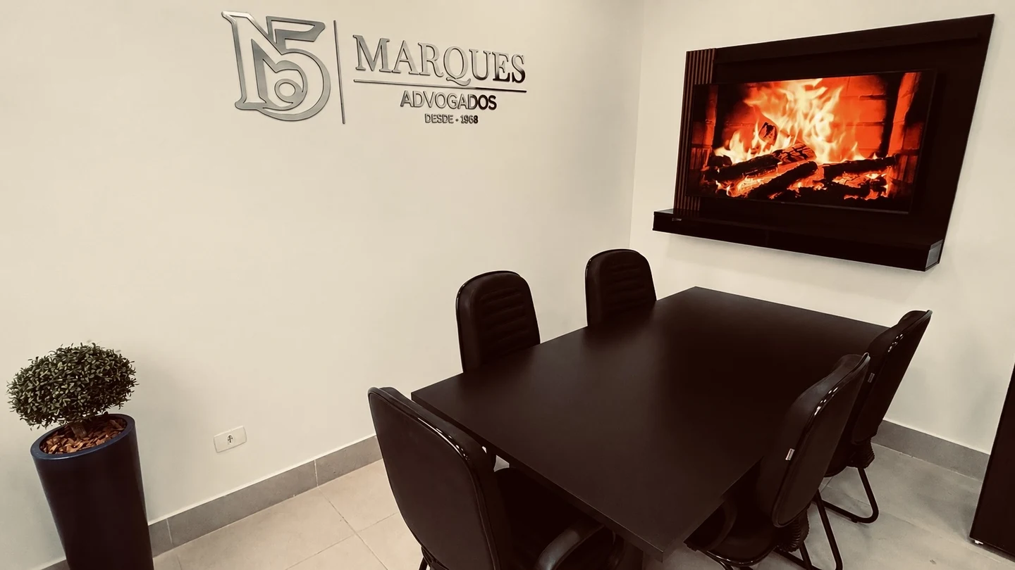 Sala de reunião do Marques Advogados, com mesa preta para seis lugares, cadeiras estofadas, televisão na parede e o logotipo do escritório aplicado na parede clara